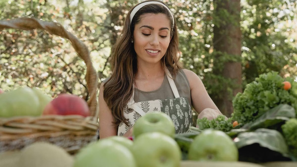 Woman with headband picking fresh apples and vegetables in a basket, smiling outdoors.